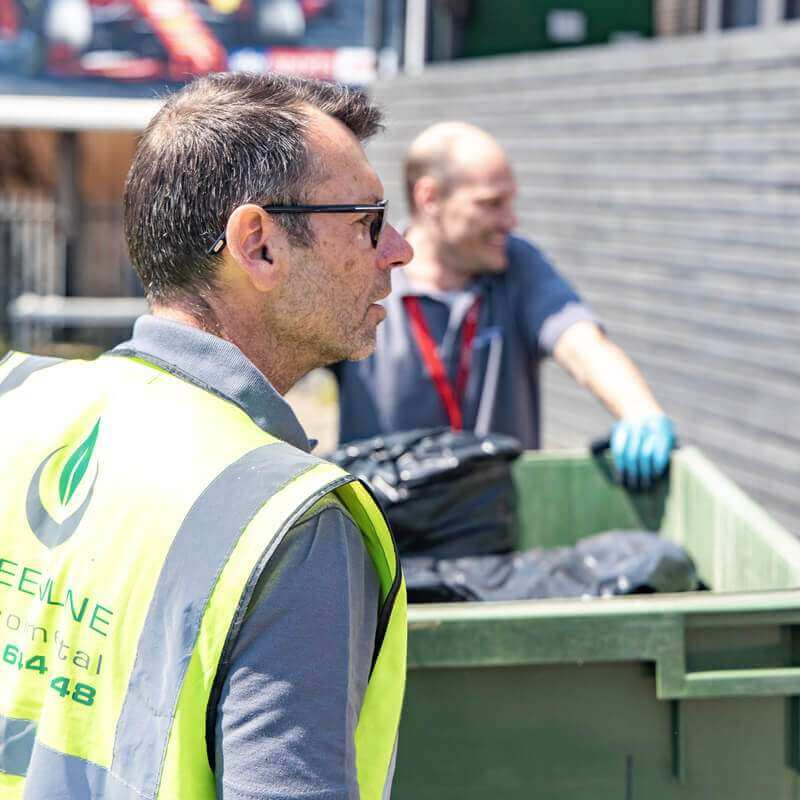 A man with safety glasses and a yellow vest observes a garbage bin, highlighting his role in waste management.
